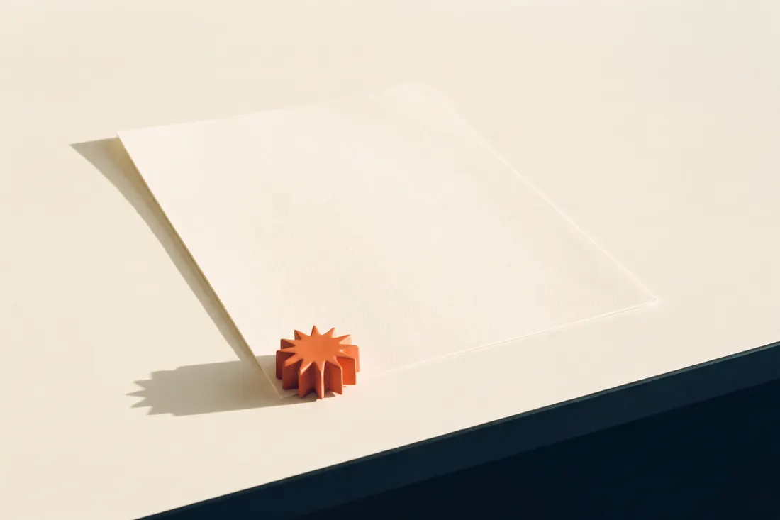 Ein einzelnes Blatt auf einem leeren Schreibtisch im Morgenlicht, beschwert von einem kleinen orangen Stern.